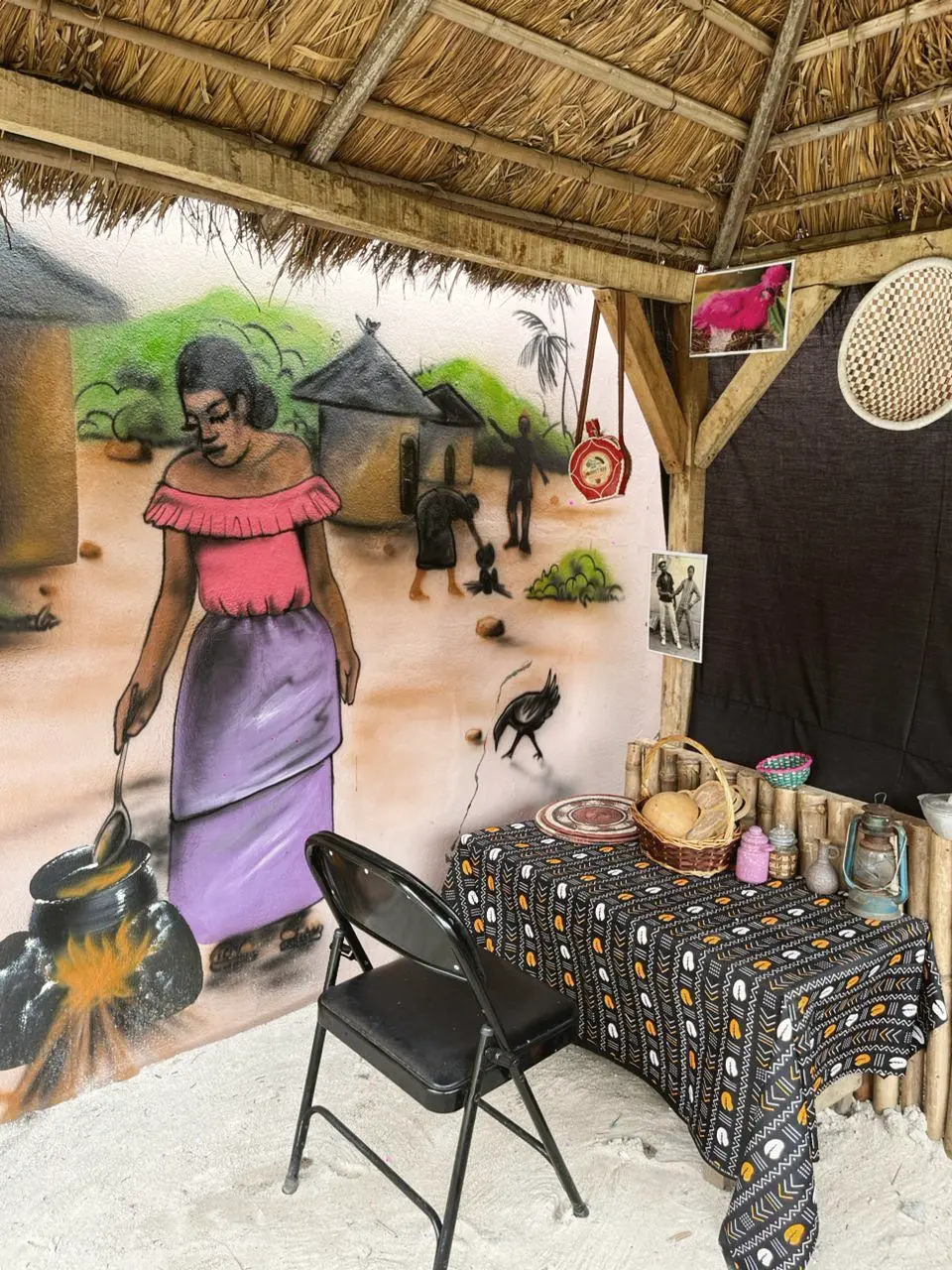 Table nappée de bogolan sous une paillote, devant une fresque de scène de village